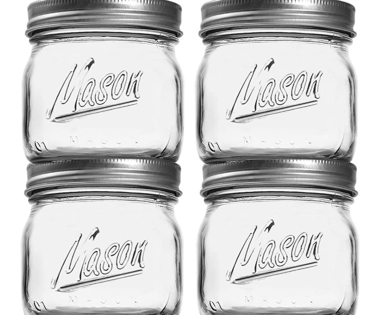 Four clear, empty Mason jars with metal lids are shown in a 2x2 arrangement. The word "Mason" is embossed on the front of each jar. These versatile jars can easily be transformed into charming mason jar luminaries for any occasion.